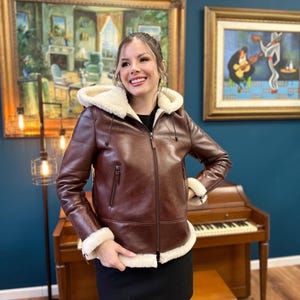 Peut inclure: Une veste en cuir marron avec une doublure en peau de mouton blanche et une capuche. La veste a une fermeture &eacute;clair et deux poches sur le devant. Le mannequin porte une jupe noire et se tient devant un piano.