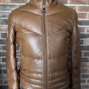 Puede incluir: Chaqueta acolchada de cuero marrón con cuello alto y cremallera completa. La chaqueta tiene un diseño acolchado y dos bolsillos con cremallera. La chaqueta se exhibe en un maniquí contra una pared de ladrillos.