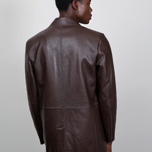 May include: A brown leather jacket with a long cut. The jacket has a simple design with a single button closure and a collar.