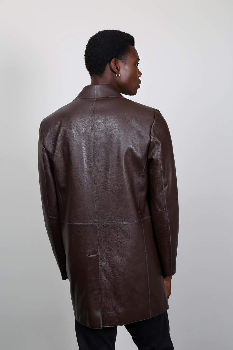 May include: A brown leather jacket with a long cut. The jacket has a simple design with a single button closure and a collar.