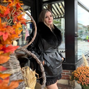 May include: A black leather jacket with a black fur collar and a belt. The jacket has a button closure and two pockets. The model is wearing a black skirt and is standing in front of a store.