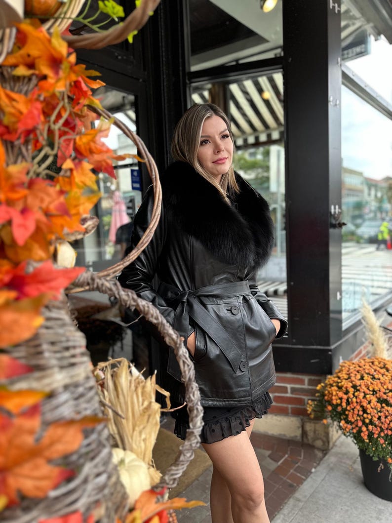 May include: A black leather jacket with a black fur collar and a belt. The jacket has a button closure and two pockets. The model is wearing a black skirt and is standing in front of a store.