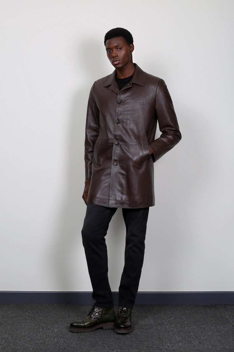 May include: A brown leather jacket with a button-up front and long sleeves. The jacket is worn by a person with dark skin and black hair. The person is wearing black pants and dark green boots.