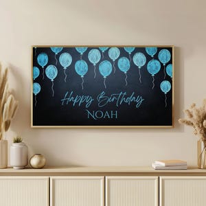 May include: A black chalkboard with a gold frame features a blue and white balloon design with the text "Happy Birthday NOAH".