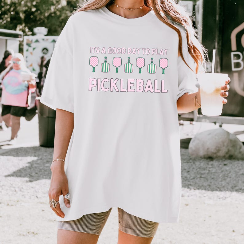 May include: White oversized t-shirt with a pink graphic that says "It's a good day to play pickleball" with a row of green and pink pickleball paddles.