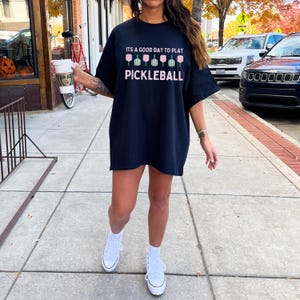 May include: A black t-shirt with the text "It's a good day to play pickleball" and a graphic of pink pickleball paddles. The person wearing the shirt is standing on a sidewalk.