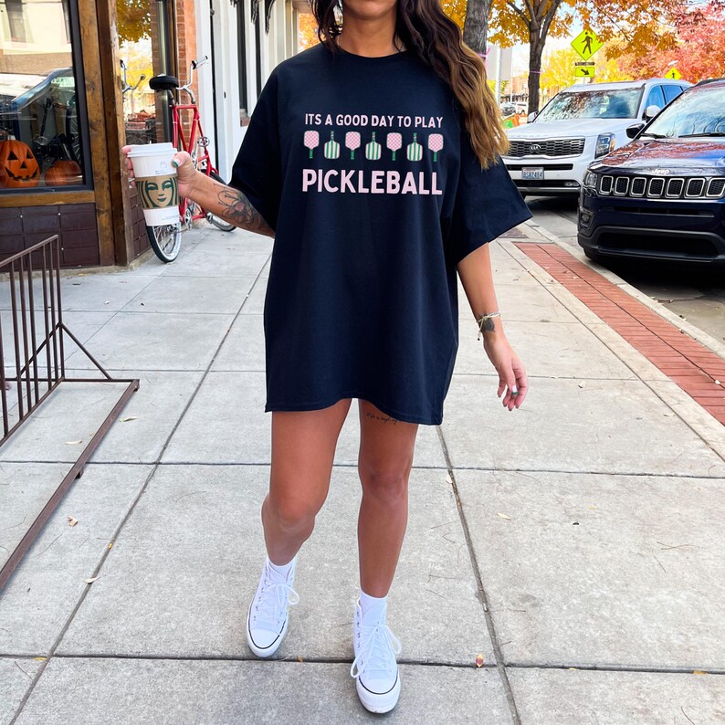 May include: A black t-shirt with the text "It's a good day to play pickleball" and a graphic of pink pickleball paddles. The person wearing the shirt is standing on a sidewalk.