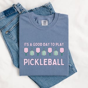 May include: A blue t-shirt with the text "It's a good day to play pickleball" in pink letters. The shirt features a row of seven pink and green pickleball paddles.