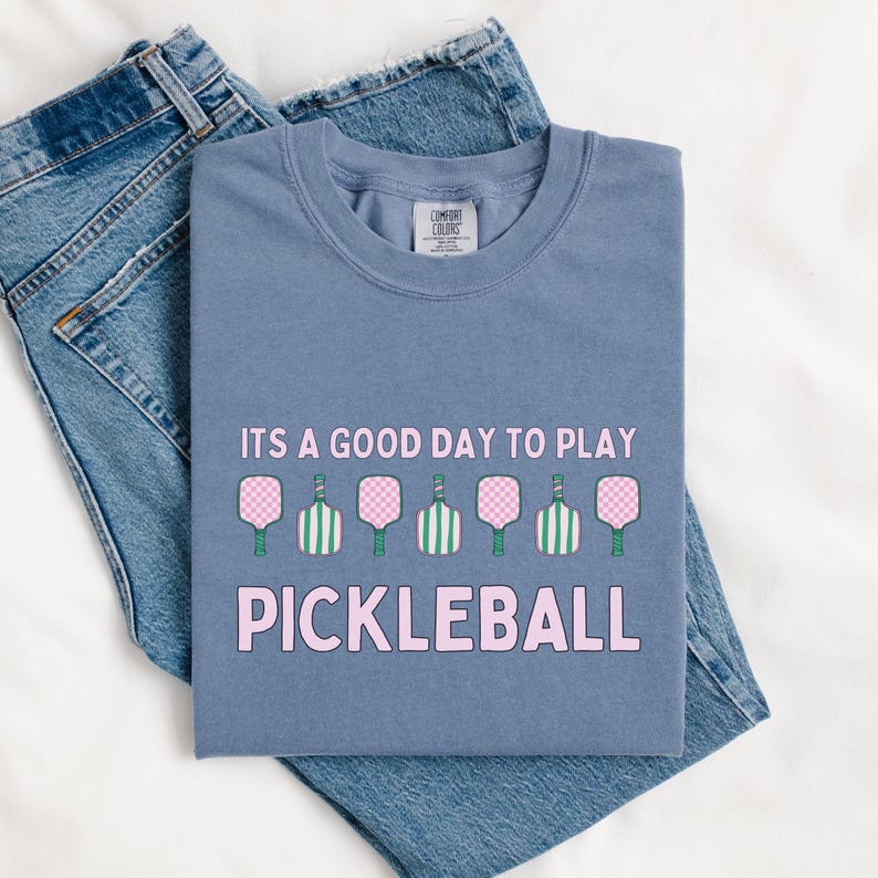 May include: A blue t-shirt with the text "It's a good day to play pickleball" in pink letters. The shirt features a row of seven pink and green pickleball paddles.
