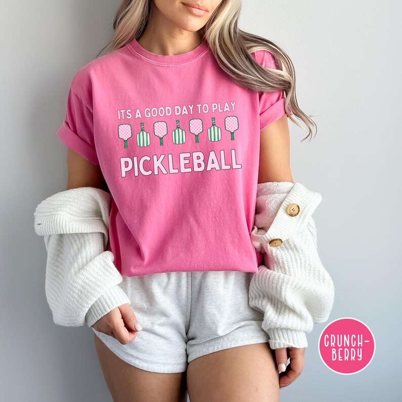May include: A pink t-shirt with the text "It's a good day to play pickleball" and a row of pickleball paddles in green and pink.
