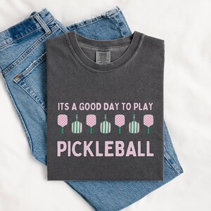 May include: A dark gray t-shirt with the text "It's a good day to play pickleball" in pink lettering. The shirt features a row of pink and green pickleball paddles.