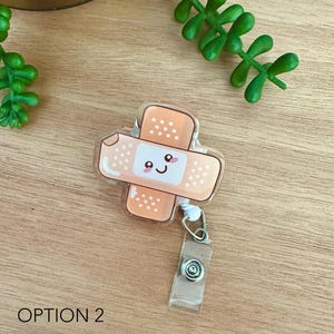 May include: A clear plastic badge holder with a cute cartoon bandage design. The bandage is orange and has a smiling face with pink cheeks. The badge holder has a retractable cord and a clip.
