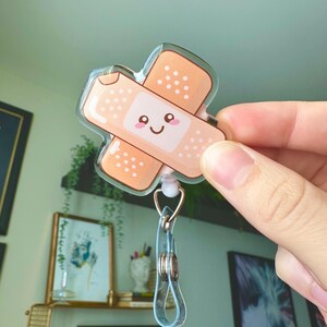 May include: A clear acrylic badge holder with a cute cartoon bandage design. The bandage has a smiling face and is in shades of brown and white.