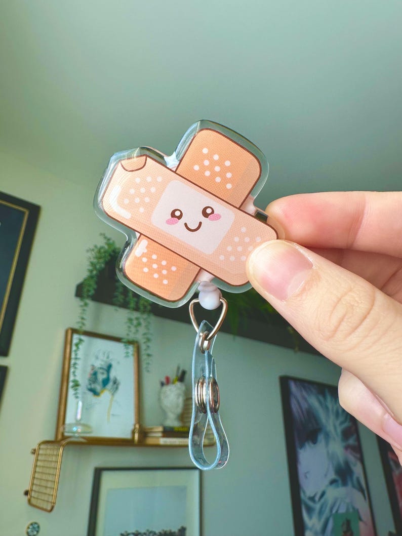 May include: A clear acrylic badge holder with a cute cartoon bandage design. The bandage has a smiling face and is in shades of brown and white.