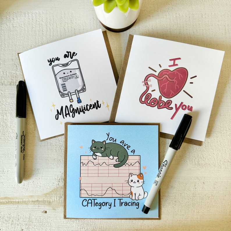 May include: Three greeting cards with humorous illustrations and text. The first card features a cartoon IV bag with the text "You are Magnificent". The second card features a cartoon heart with the text "I lobe you". The third card features a cartoon cat on a heart rate monitor with the text "You are a Category I Tracing".