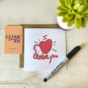 May include: A white card with a red anatomical heart illustration and the text "I lobe you". The card is on a brown surface with a black marker and a small pink card with the text "The Art Clinic RN @the_artclinic" on it.