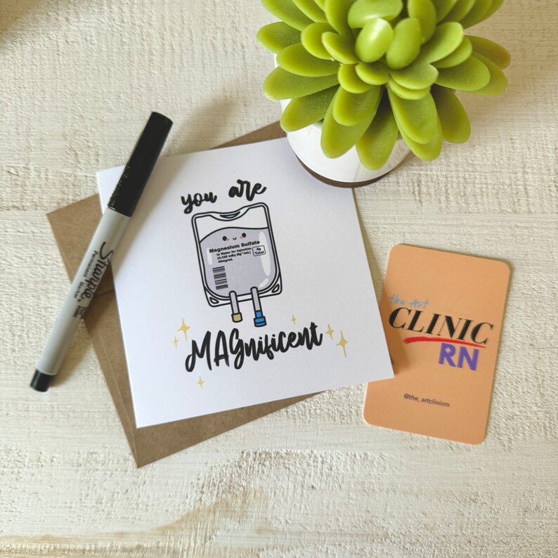 May include: A white card with a cartoon illustration of an IV bag with the text "you are MAGnificent". The card is on a brown surface with a black marker and a small orange card with the text "the art clinic RN" on it.