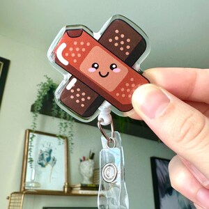 May include: A clear plastic badge holder with a cute cartoon band-aid design. The band-aid is red and brown with a smiling face.