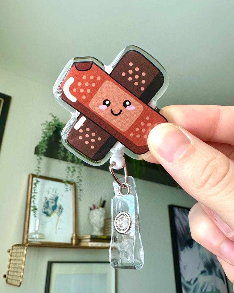 May include: A clear plastic badge holder with a cute cartoon band-aid design. The band-aid is red and brown with a smiling face.