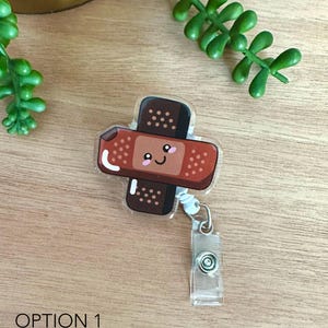 May include: A brown and white cartoon bandaid with a smiling face on a clear plastic retractable badge holder. The badge holder has a silver clip and a clear plastic strap.