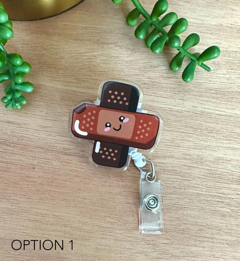 May include: A brown and white cartoon bandaid with a smiling face on a clear plastic retractable badge holder. The badge holder has a silver clip and a clear plastic strap.
