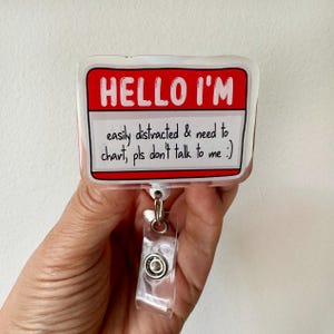 May include: A rectangular badge with the text "HELLO I'M" in white on a red background. Below, it reads "easily distracted & need to chart, pls don't talk to me :)". The badge is attached to a clear plastic retractable reel.