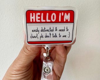 May include: A rectangular badge with the text "HELLO I'M" in white on a red background. Below, it reads "easily distracted & need to chart, pls don't talk to me :)". The badge is attached to a clear plastic retractable reel.