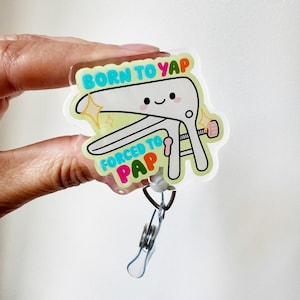 May include: A clear acrylic badge reel featuring a cartoon illustration of a white medical speculum with a smiling face. The text BORN TO YAP FORCED TO PAP is printed in colorful letters. The design includes yellow star accents on a light green background.