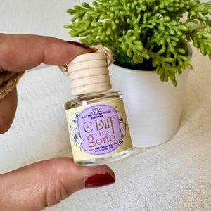 May include: A small, clear glass bottle with a wooden cap and a label that reads "C. Diff be gone." The label is purple and yellow with floral accents. The bottle is held in front of a small potted plant.