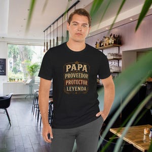 May include: A black short-sleeved t-shirt featuring a graphic design with the text PAPA, PROVEEDOR, PROTECTOR, LEYENDA in a stacked, distressed font style, accented with a small crown icon at the top and decorative border elements.