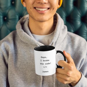 May include: White coffee mug with a black interior and handle. The mug has the text "Oops, I broke the code! </> James" printed in black. The mug is held by a person wearing a gray hoodie.