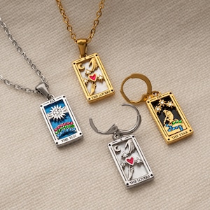 May include: Four tarot card pendants and earrings. The pendants are rectangular with gold or silver frames. The designs include a sun, a heart, and a figure under a starry sky. The earrings are gold and silver with a tarot card design.
