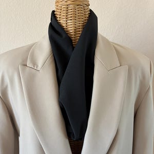 May include: A beige blazer with a black scarf draped around a mannequin's neck. The mannequin has a woven wicker head and a wooden top. The blazer has a classic lapel design.