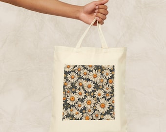 Op de afbeelding: Een witte canvas tote bag met een bloemenprint. De print heeft een patroon van witte madeliefjes met gele centers op een donkere achtergrond.