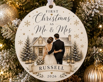 Op de afbeelding: Een ronde witte kerstornament met een koppel voor een huis met de tekst First Christmas as Mr & Mrs, de naam RUSSELL op een banner en het jaartal 2026, versierd met gouden sneeuwvlokken, sterren en dennenbomen.
