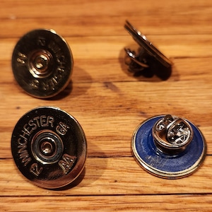 May include: Three gold-colored pins with different designs. One pin features a blue and gold design with a pin back. The other two pins feature a gold-colored shotgun shell with the text "12 Winchester GA AA" printed on the shell.