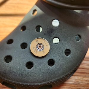 May include: Close-up of a dark gray clog shoe with a gold-colored bullet-shaped charm. The charm has the text "12 GA" and "MIN" engraved on it. The shoe has several round holes.