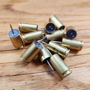 May include: A group of brass bullet casings with push pins inserted into the bottom. The casings are arranged in a pile on a wooden surface.