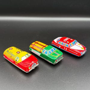 May include: Three vintage tin toy vehicles including a red and yellow Mobilgas tanker truck, a green and yellow Lumber Trax truck, and a red and white Chief fire department car. Each vehicle features painted details, windows, and small driver figures.