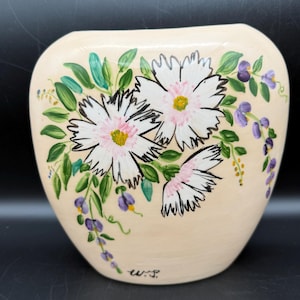 May include: Cream-colored ceramic vase with a wide, rounded shape. The vase is hand-painted with white daisies, green leaves, and purple wisteria-like flowers. The artist's initials, "w.s.", are visible near the base.