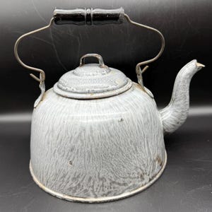 May include: A vintage gray and white speckled enamelware kettle with a black wooden handle. The kettle has a spout and a lid with a small handle. The handle is made of metal. The kettle is a classic kitchenware item.