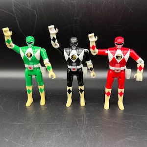 May include: Three Power Ranger action figures in a row. The figures are in green, black, and red. Each figure has a white helmet, a diamond-shaped emblem on the chest, and matching colored boots. The figures are standing with their arms raised.