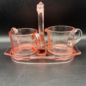 May include: A vintage pink glass creamer and sugar set. The set includes a creamer, a sugar bowl, and a tray with a handle. The pieces are made of clear pink glass and have a simple, elegant design. The set is likely from the mid-20th century.
