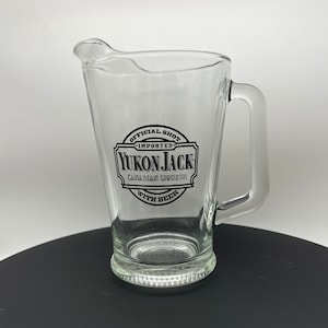 May include: A clear glass pitcher with a handle. The pitcher has a black and white label that says "Official Shot - Imported - Yukon Jack - Canadian Liqueur - With Beer".