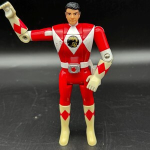 May include: A Power Rangers action figure in a red and white uniform with a black and gold emblem on the chest. The figure has articulated arms and legs, and is wearing white gloves and boots. The figure is standing on a black surface.