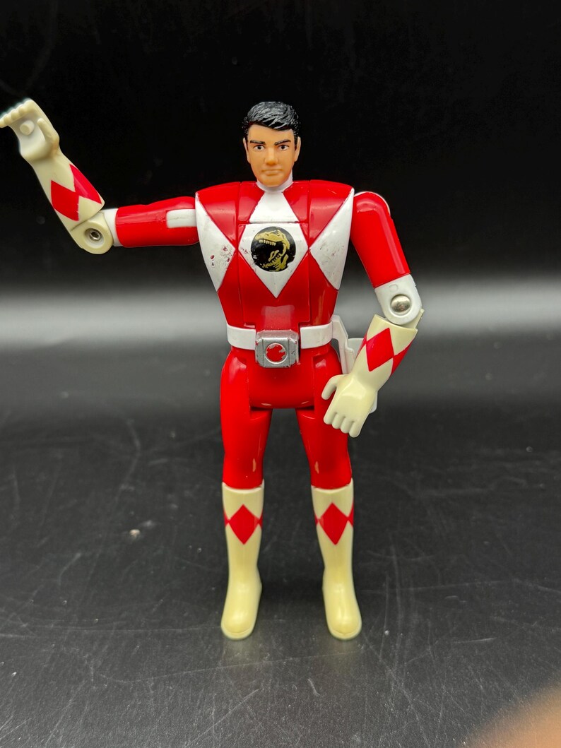 May include: A Power Rangers action figure in a red and white uniform with a black and gold emblem on the chest. The figure has articulated arms and legs, and is wearing white gloves and boots. The figure is standing on a black surface.