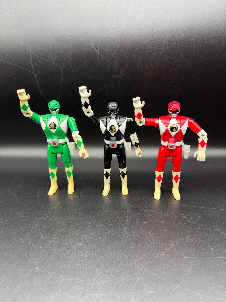 May include: Three Power Ranger action figures in a row. The figures are in green, black, and red. Each figure has a white helmet, a diamond-shaped emblem on the chest, and matching colored boots. The figures are standing with their arms raised.