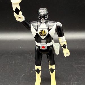 May include: Black Power Ranger action figure with white and black diamond accents. The figure has a black helmet and uniform with a white diamond on the chest. The figure is wearing yellow boots and has a white hand raised.