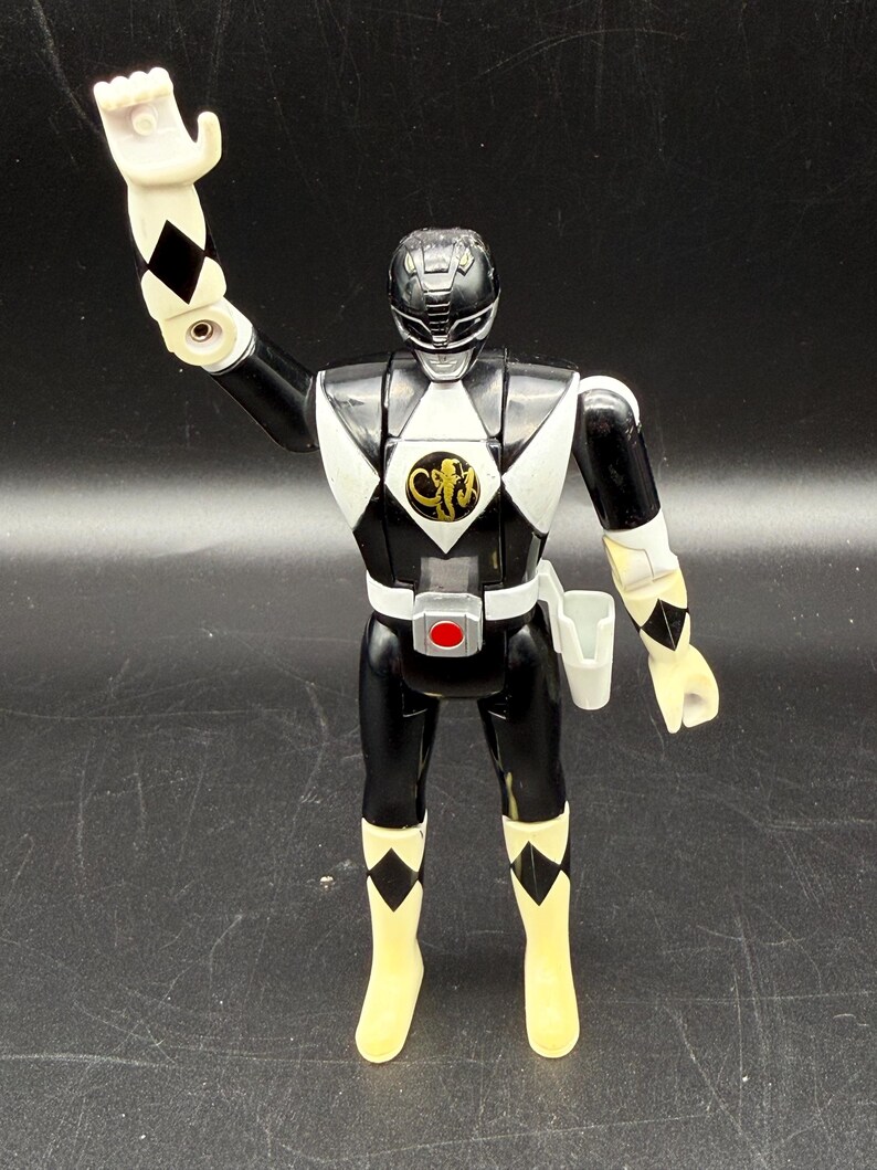 May include: Black Power Ranger action figure with white and black diamond accents. The figure has a black helmet and uniform with a white diamond on the chest. The figure is wearing yellow boots and has a white hand raised.