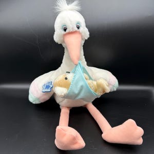 May include: A plush stork toy with a white body, pink beak, legs, and feet. The stork carries a baby in a light blue sling. The wings have pastel pink and green accents. A blue patch with a hand graphic is on the wing.